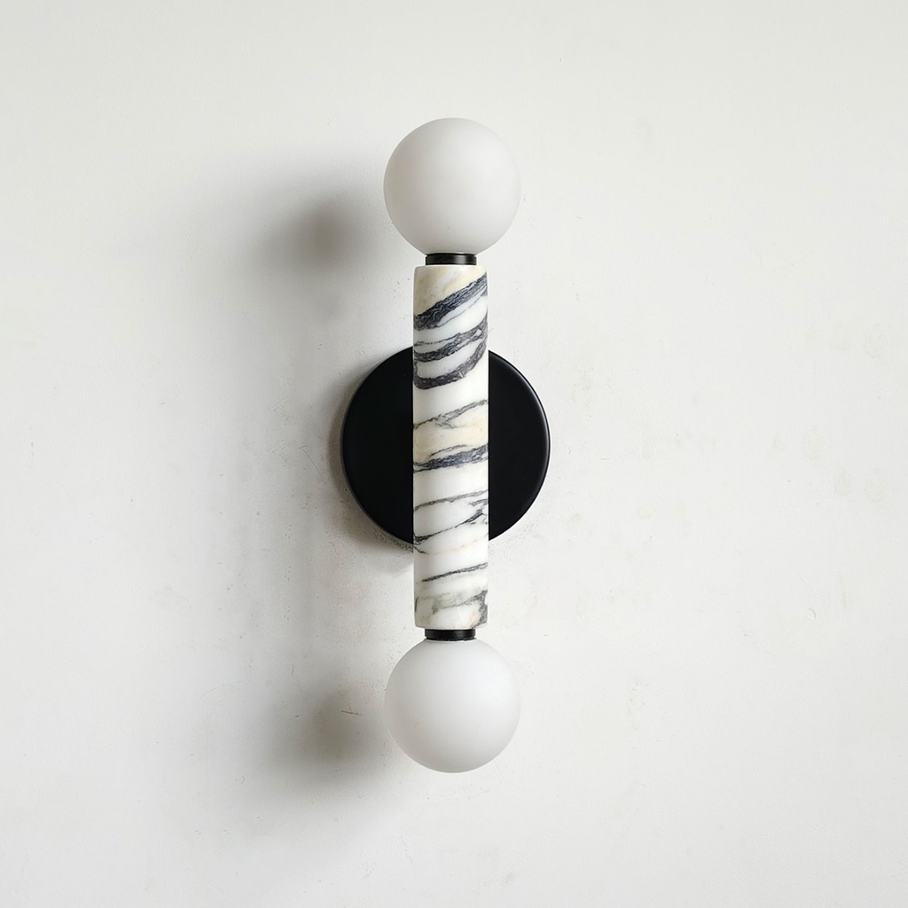 A modern wall-mounted sconce with two white spherical shades, a central cylindrical Calacatta marble column with black veining, and a round black backplate on a white wall.