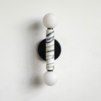 A modern wall-mounted sconce with two white spherical shades, a central cylindrical Calacatta marble column with black veining, and a round black backplate on a white wall.