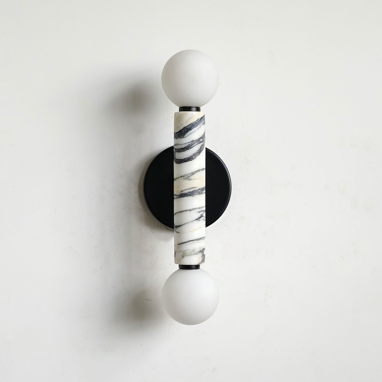 A modern wall-mounted sconce with two white spherical shades, a central cylindrical Calacatta marble column with black veining, and a round black backplate on a white wall.