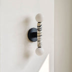 A modern wall-mounted light fixture with two white spherical shades and a central cylindrical marble column featuring black veining, mounted on a black circular backplate against a white wall.