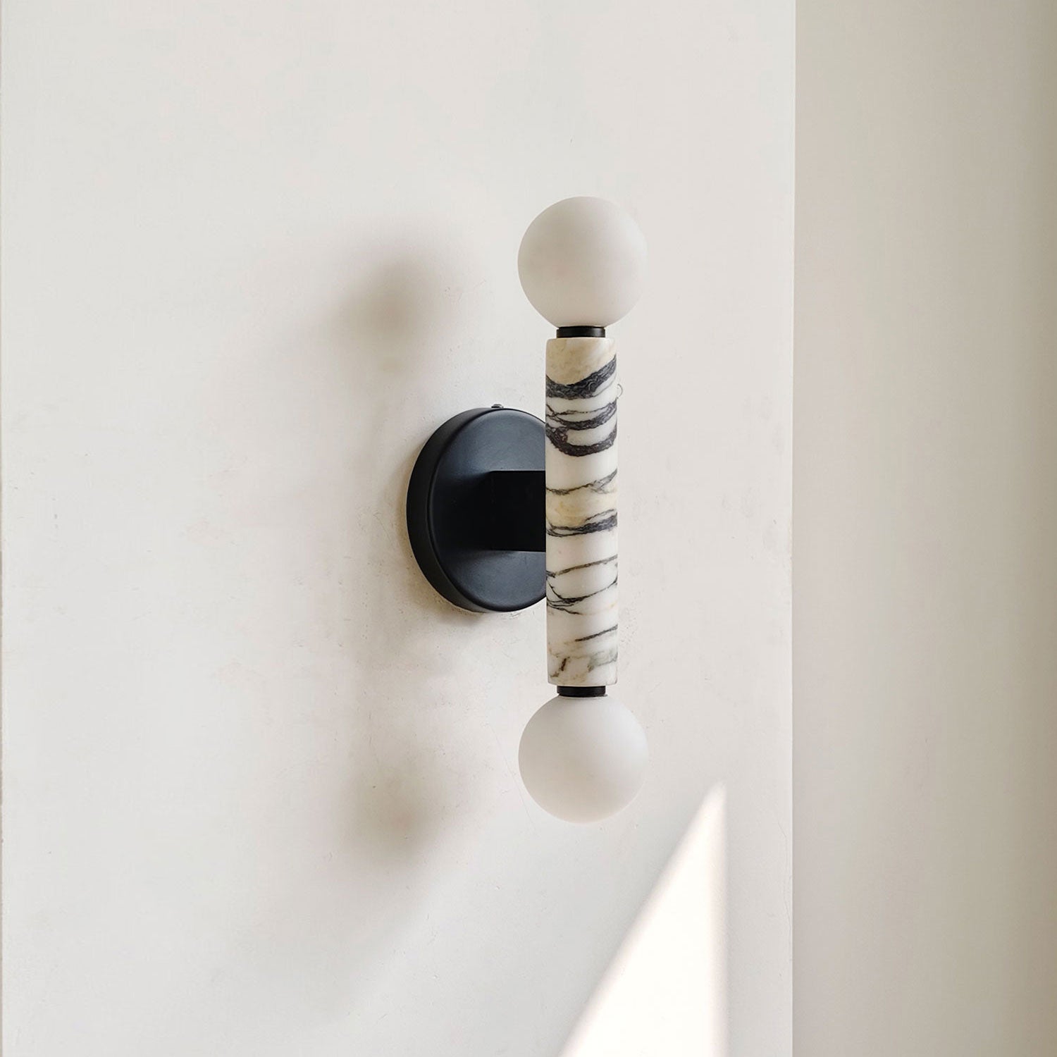 A modern wall-mounted light fixture with two white spherical shades and a central cylindrical marble column featuring black veining, mounted on a black circular backplate against a white wall.