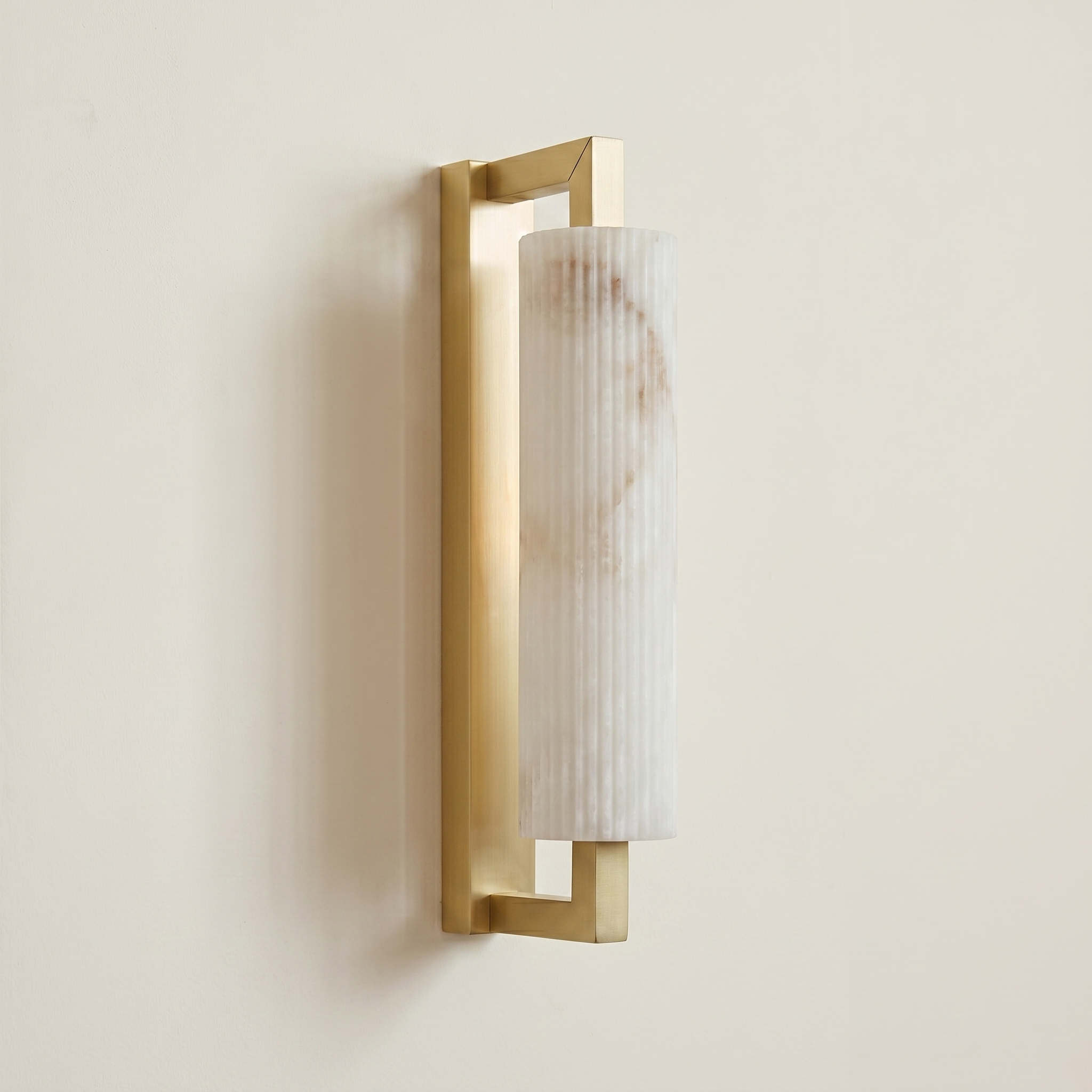 Pilaster Alabaster Cylinder light fixture with a tall, cylindrical alabaster shade and a brushed brass metal frame, shown illuminated against a plain off-white wall. | IINK Interiors