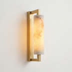 Pilaster Alabaster Cylinder light fixture with a tall, cylindrical alabaster shade and a brushed brass metal frame, shown illuminated against a plain off-white wall. | IINK Interiors