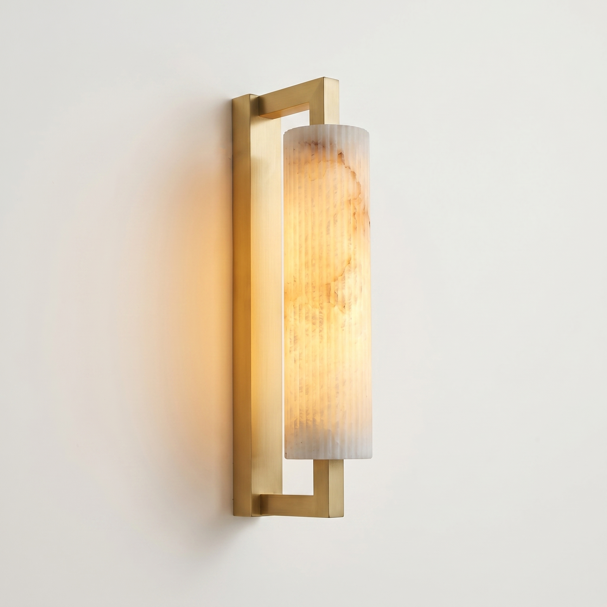 Pilaster Alabaster Cylinder light fixture with a tall, cylindrical alabaster shade and a brushed brass metal frame, shown illuminated against a plain off-white wall. | IINK Interiors