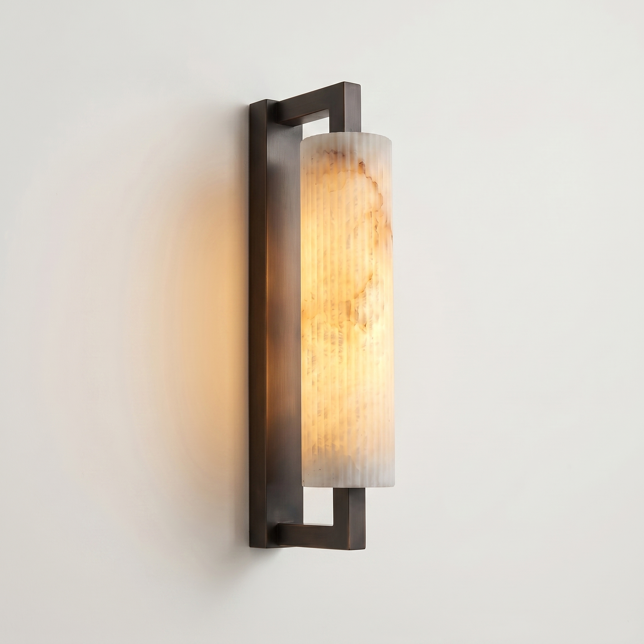 Pilaster Alabaster Cylinder light fixture with a tall, cylindrical alabaster shade and a brushed bronze metal frame, shown illuminated against a plain off-white wall. | IINK Interiors