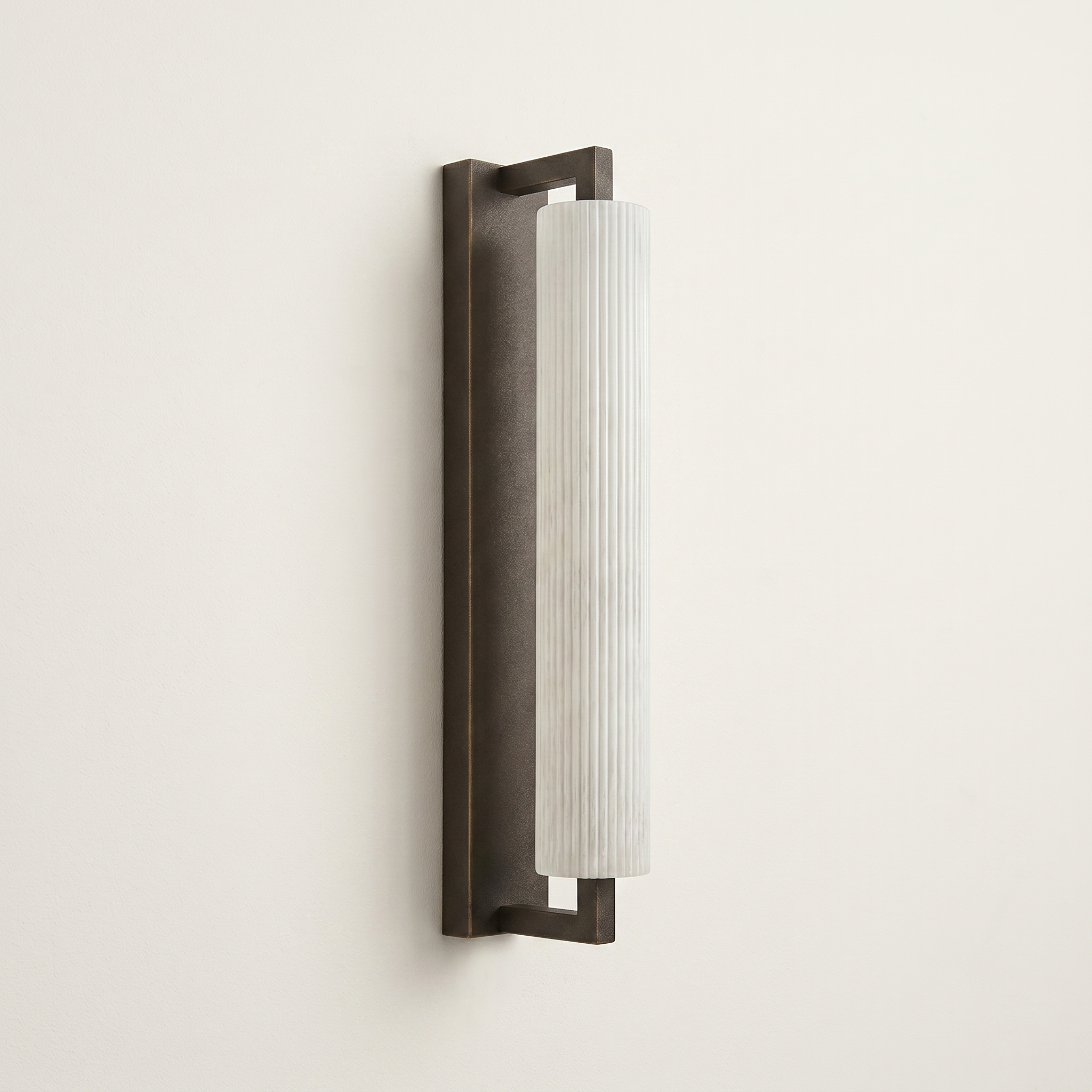 Pilaster Alabaster Cylinder light fixture with a tall, cylindrical alabaster shade and a brushed bronze metal frame, shown illuminated against a plain off-white wall. | IINK Interiors