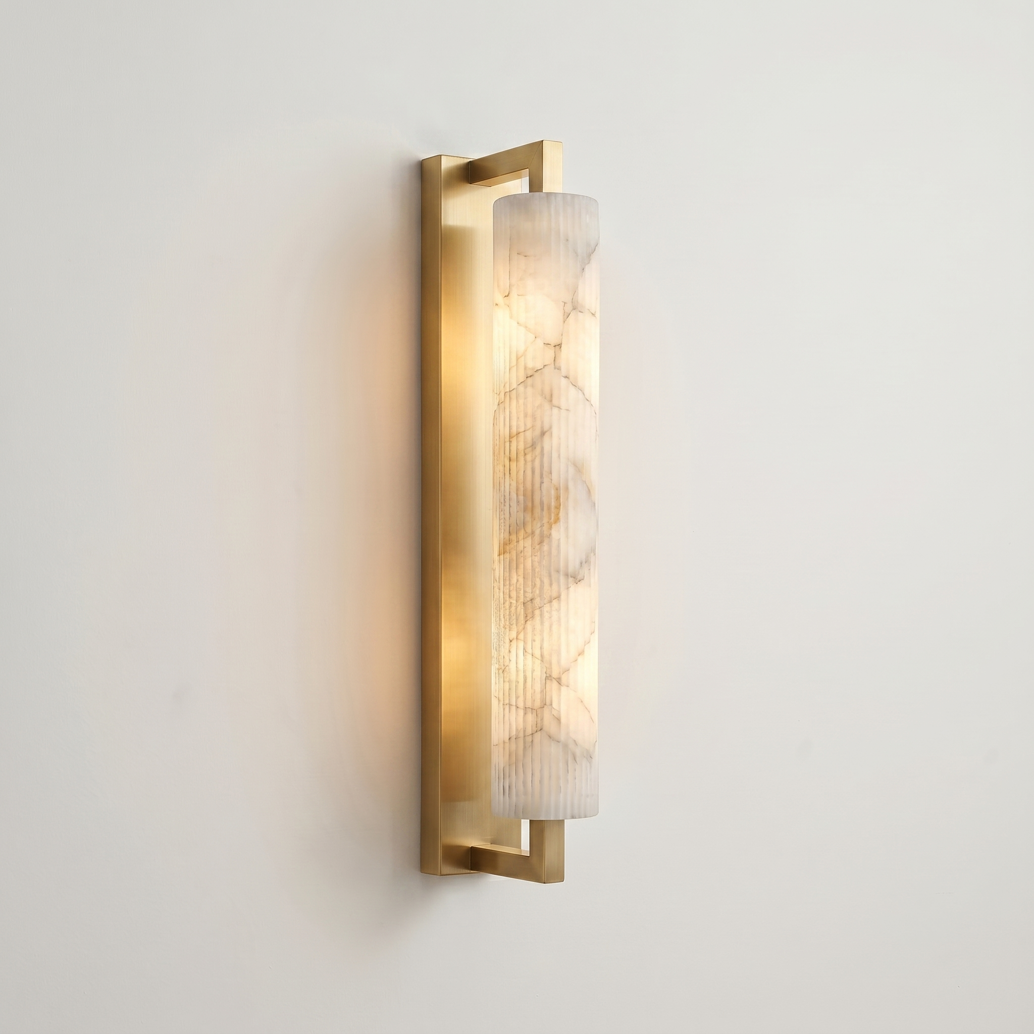 Pilaster Alabaster Cylinder light fixture with a tall, cylindrical alabaster shade and a brushed brass metal frame, shown illuminated against a plain off-white wall. | IINK Interiors