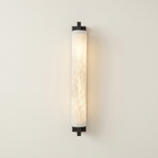 Pilaster Alabaster Cylinder light fixture with a tall, cylindrical alabaster shade and a brushed bronze metal frame, shown illuminated against a plain off-white wall. | IINK Interiors
