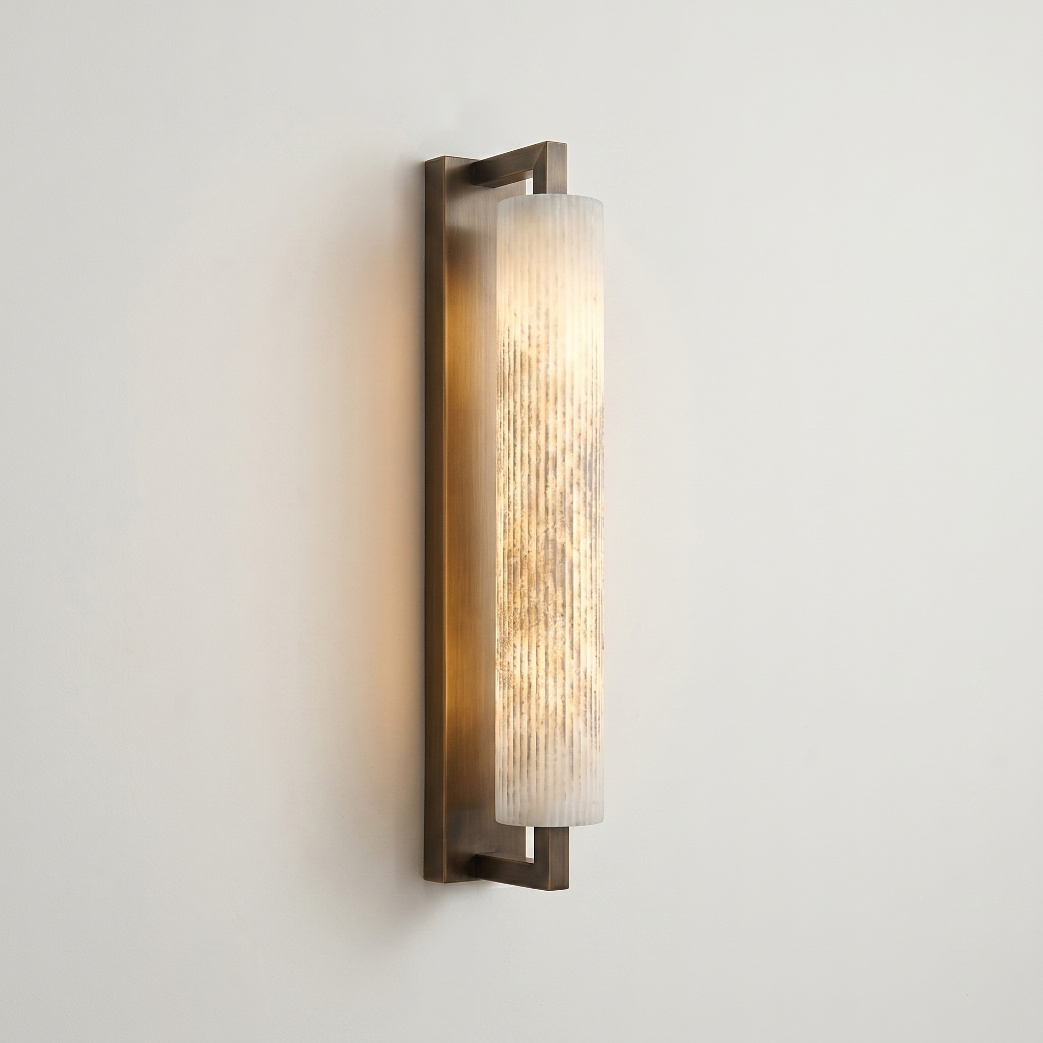 A modern wall sconce light fixture with a vertical cylindrical alabaster stone shade and a brushed bronze metal frame, shown illuminated against a plain off-white wall. | IINK Interiors