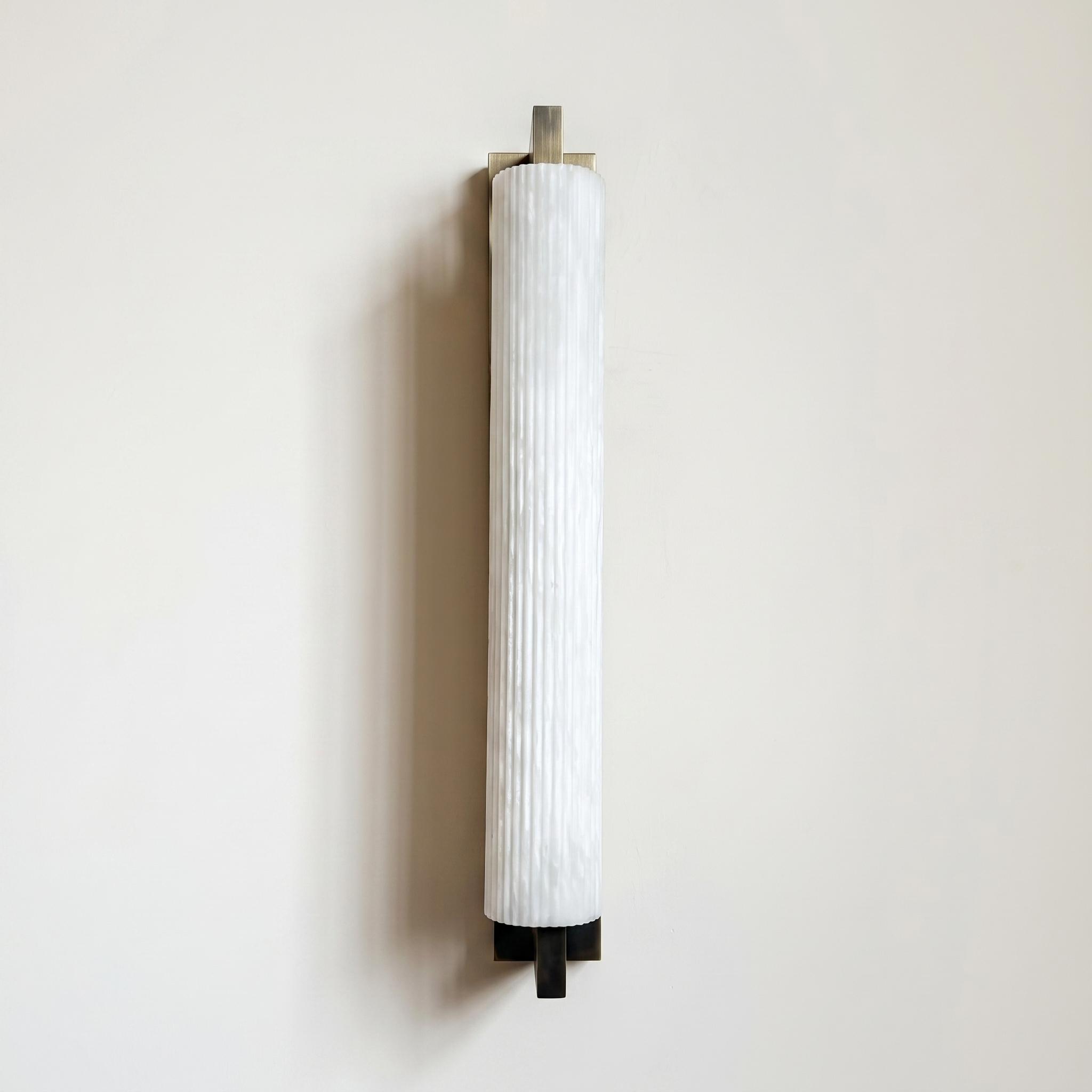 Pilaster Alabaster Cylinder light fixture with a tall, cylindrical alabaster shade and a brushed bronze metal frame, shown illuminated against a plain off-white wall. | IINK Interiors